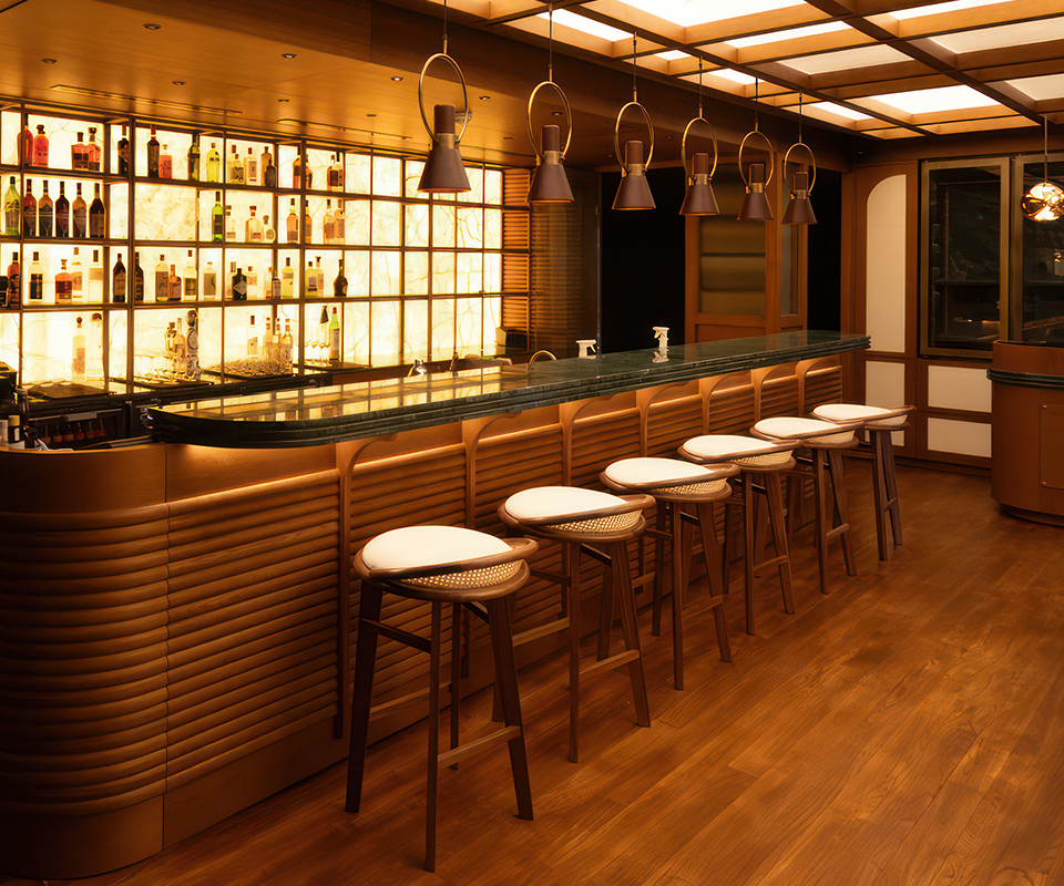 Beefbar Restaurant, Luxembourg A long row of Brummell bar stools with a combination of dark wood, light straw weave (cannage), and a white upholstered seat. The stools are positioned at a green marble-top bar with vertical wood cladding. The atmosphere is cozy and warm, illuminated by a back-lit bottle wall and several bronze suspension lamps at Beefbar Restaurant, Luxembourg.