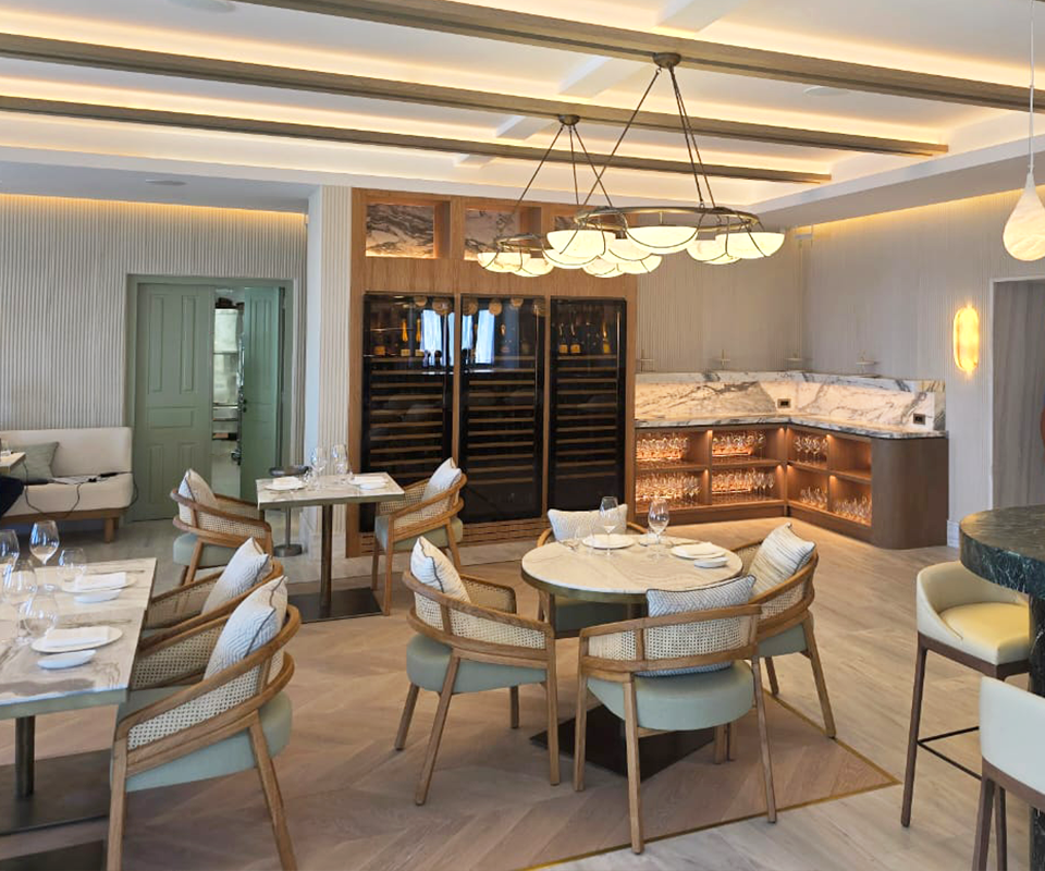 Elegant dining room design at Blu Restaurant in Santorini, Greece, showcasing light spencer dining chairs with cane backrests by Wood Tailors Club around marble-top tables under a contemporary chandelier.