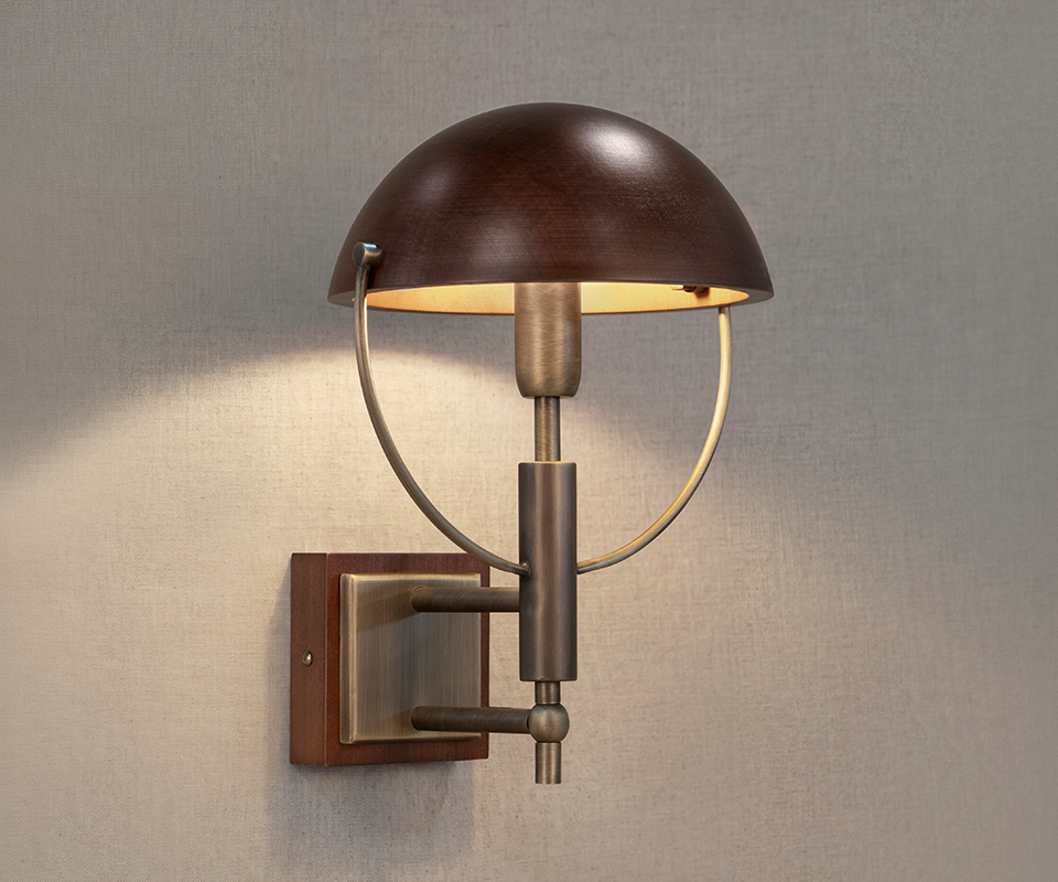 Faraday Wall Lamp Faraday Wall Lamp by Wood Tailors Club switched on against a beige fabric-textured wall, casting a warm glow through its smoked walnut shade and aged brushed brass structure.