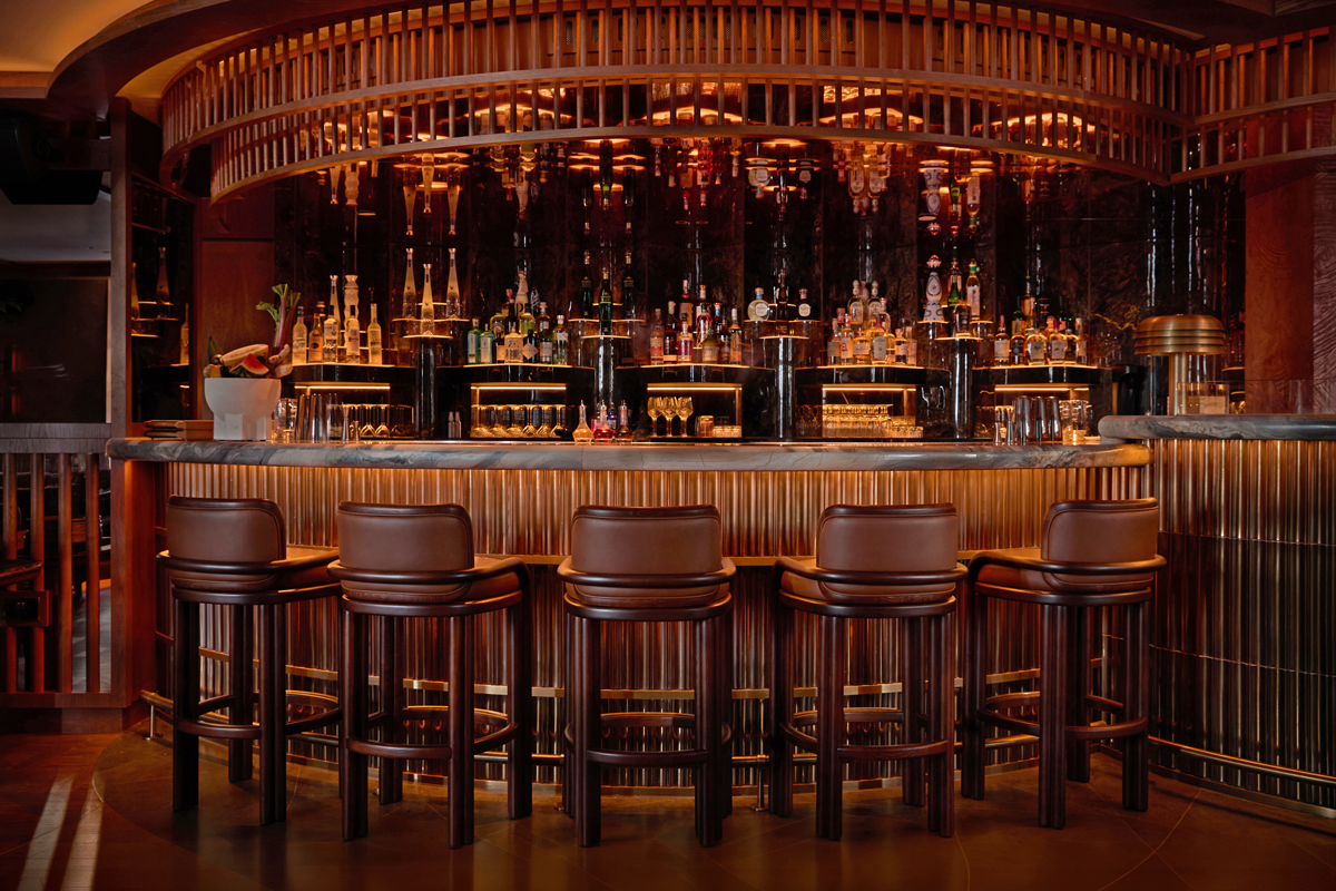 Close-up photograph of the opulent bar counter at MA/NA Mayfair. The circular bar features a veined marble top and a base clad in fluted golden metallic panels that catch the embedded lighting. Lined up in front of the counter are five high bar stools in brown leather with dark wood frames, supplied by Emotional Brands. In the background, the richly illuminated bar shelves display premium spirit bottles against a dark, mirrored backdrop under a curved wooden slat canopy.