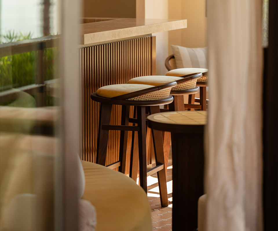 Pool House by Studio Quentin, Monte-Carlo, Monaco A row of Brummell bar stools with a combination of dark wood, light rattan, and a beige upholstered seat. The stools are lined up at a bar counter made of vertical wood strips. The perspective is slightly restricted, as if through an open door, with natural light coming from the left at Pool House by Studio Quentin, Monte-Carlo, Monaco.