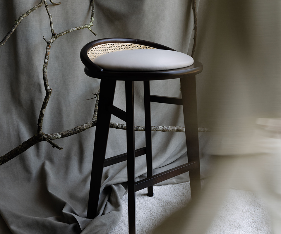 Brummell Bar Stool Brummell bar stool in black stained ashwood featuring a light grey upholstered round seat and a curved cane backrest, styled in a minimalist studio setting with neutral fabric backdrop and tree branches.