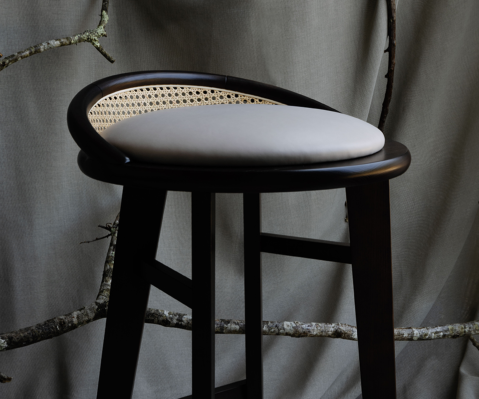 Brummell Bar Stool Close-up view of the Brummell bar stool in black stained ashwood, highlighting the light grey cushion seat and woven cane detail on the curved backrest against a draped fabric backdrop.