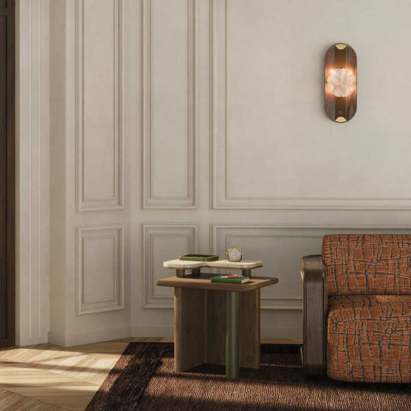 Maurier side table with a cylindrical leather-wrapped leg, wooden frame, and smooth travertine tabletop stands beside a window, paired with the Kimberley sofa upholstered in Velours Rocca by Nobilis, featuring a smoked walnut structure and rattan details. Above, the Cavendish wall lamp in smoked walnut, alabaster, leather, and aged brushed brass casts a warm glow, enriching the serene, refined ambience. This pieces are brand new arrivals in the collection.