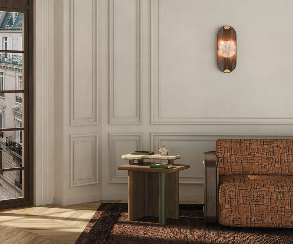 Cavendish Wall Lamp Maurier side table with a cylindrical leather-wrapped leg, wooden frame, and smooth travertine tabletop stands beside a window, paired with the Kimberley sofa upholstered in Velours Rocca by Nobilis, featuring a smoked walnut structure and rattan details. Above, the Cavendish wall lamp in smoked walnut, alabaster, leather, and aged brushed brass casts a warm glow, enriching the serene, refined ambience. This pieces are brand new arrivals in the collection.