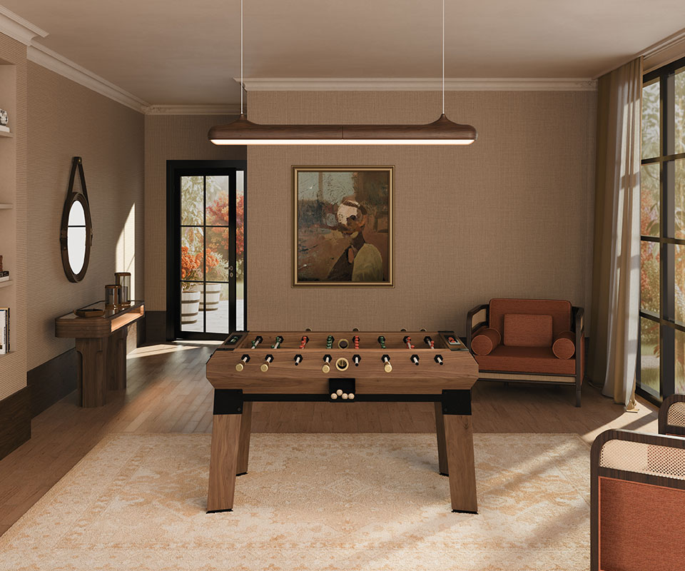 A luxurious, warm-toned game room featuring the walnut Chesterfield Foosball table at its center on a large rug, illuminated by the linear Bruce suspension lamp hanging above. Along the left wall stands the Melbourne console table with the round Reynolds mirror suspended over it. To the right, the Crockford armchair sits beside a large window bathed in sunlight.