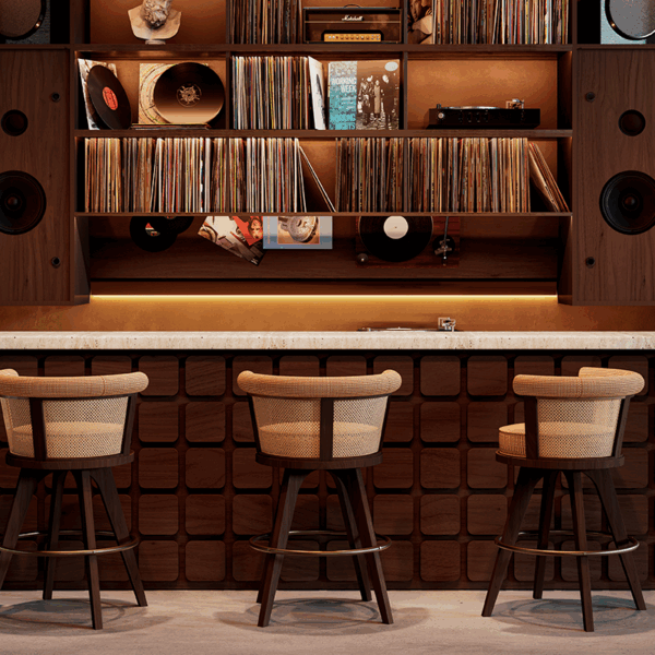 Three George Bar Chairs by Wood Tailors Club arranged in front of a mid-century modern home listening bar. The chairs feature dark walnut wood legs, a circular brass footrest, and a curved backrest with woven rattan detailing and beige upholstery. The background showcases a sophisticated wooden built-in shelving unit filled with vinyl records, large integrated speakers, and warm ambient lighting.