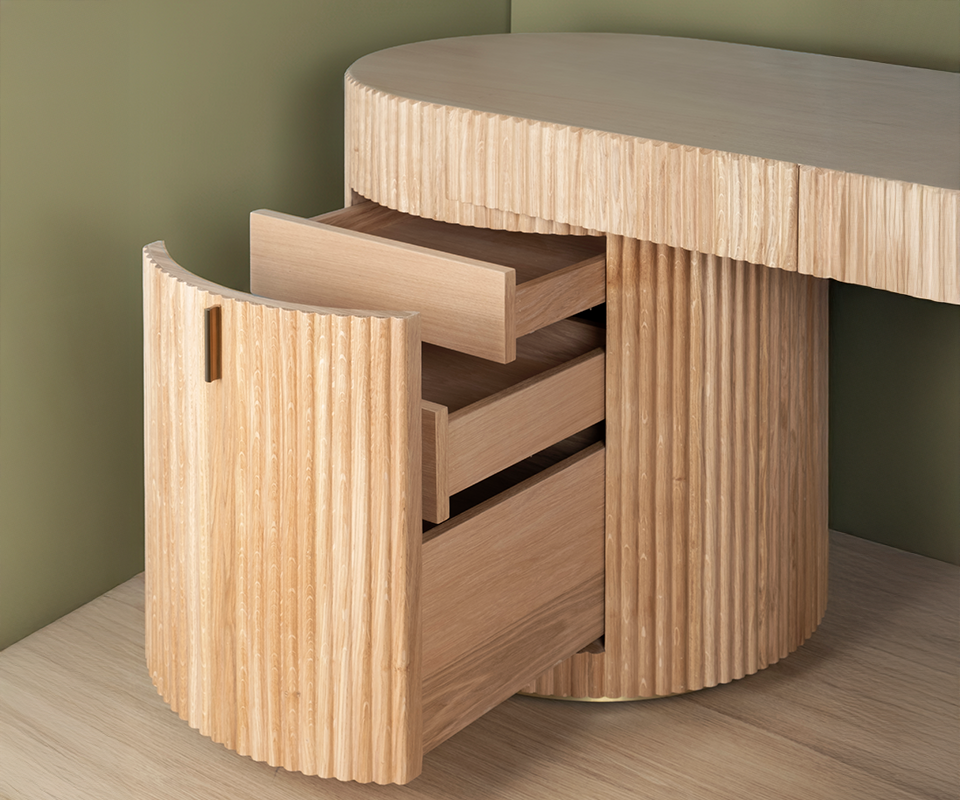 Close-up of the Gifford desk by Wood Tailors Club in oak. The curved, fluted door of the left cylindrical cabinet is open, revealing three extended inner oak drawers. A small brass pull handle details the door, with the desk resting on a light wooden floor against a soft green wall.