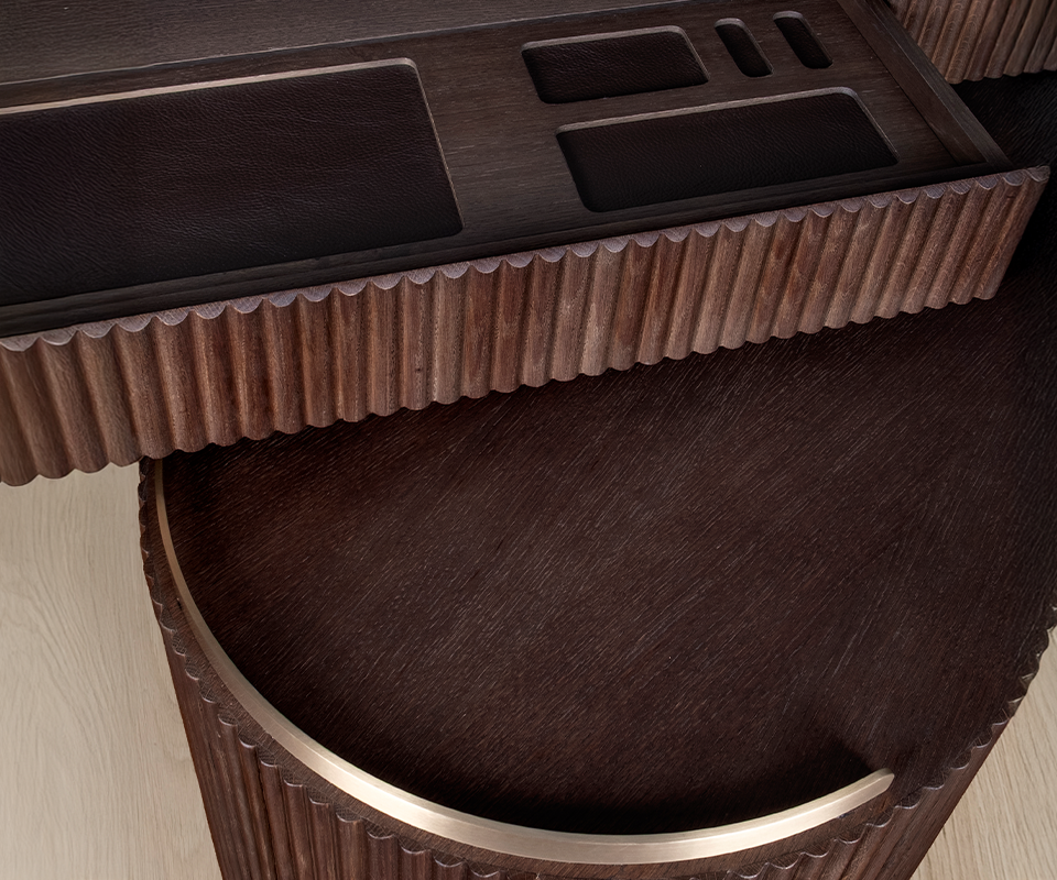 Detailed close-up of the Gifford desk by Wood Tailors Club in smoked walnut wood. An open drawer displays built-in, dark leather-lined organizational compartments. The image highlights the fluted wood texture of the drawer front and the curved edge of the lower module below, accented with a polished brass trim.