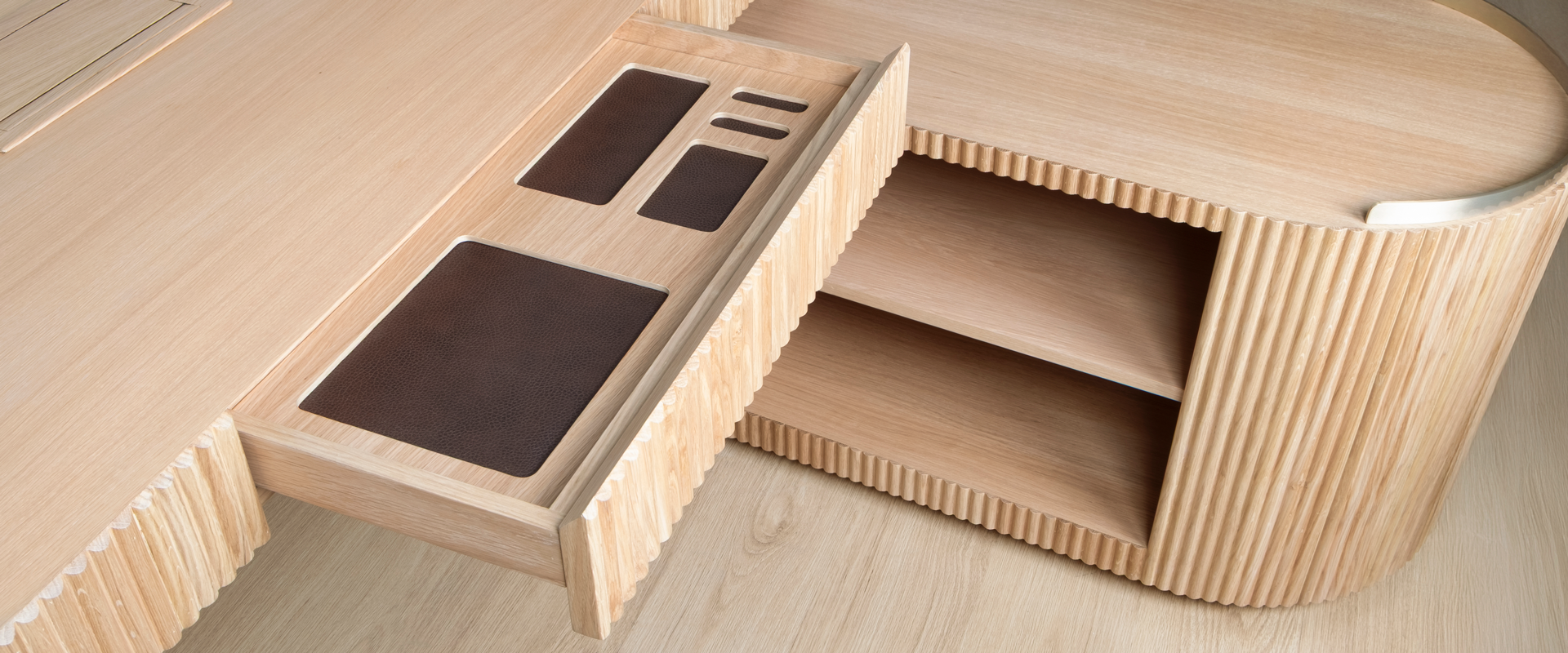 Close-up detail of the Gifford desk by Wood Tailors Club in oak. The main desktop drawer is pulled open, showcasing dark leather-lined organizer compartments. The fluted texture is prominently visible on the drawer front and on the adjacent lower oak module to the right, which reveals two open storage shelves.