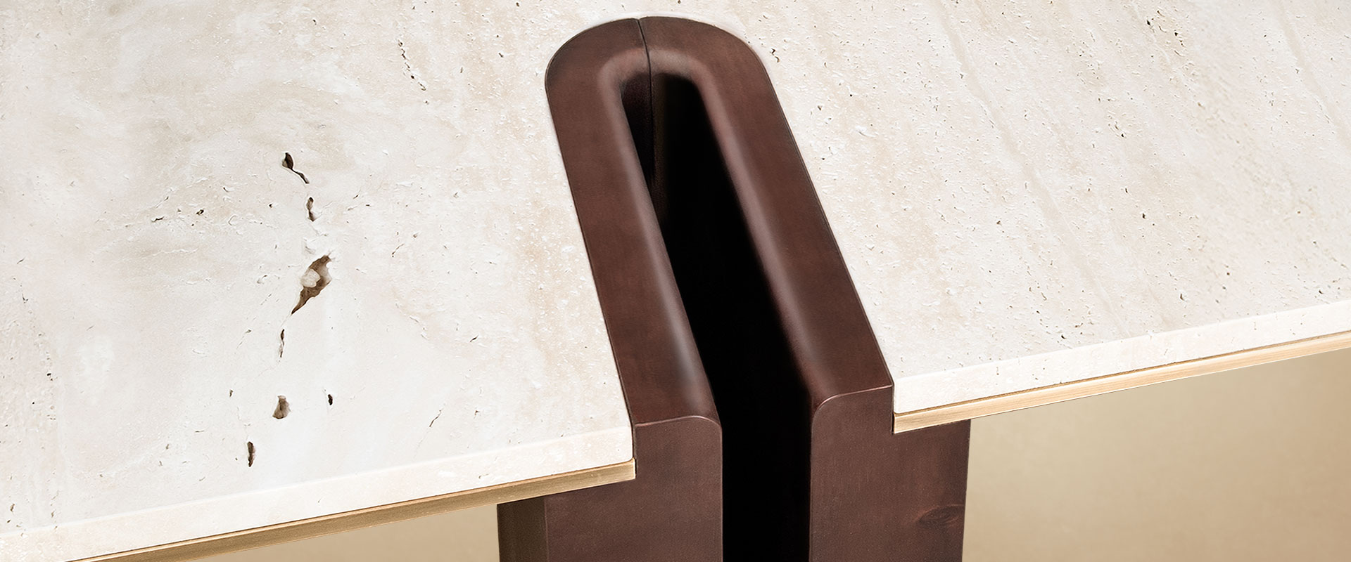 A close-up, angled detail of the Grenville Center Table's joinery. A deep, U-shaped groove in the dark smoked walnut leg interlocks with the edge of the cream travertine surface. A thin, elegant layer of brushed brass is visible as a metallic accent running horizontally between the stone top and the wooden structure.