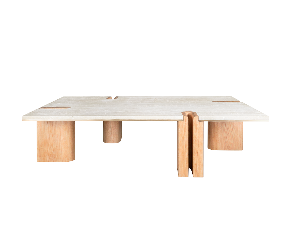 A front-facing shot of the Grenville Center Table in a lighter finish. The table features a porous cream travertine surface supported by thick, rounded light oak wood legs. The architectural U-shaped notch in the foreground leg highlights the brushed brass detailing between the wood and the stone top.