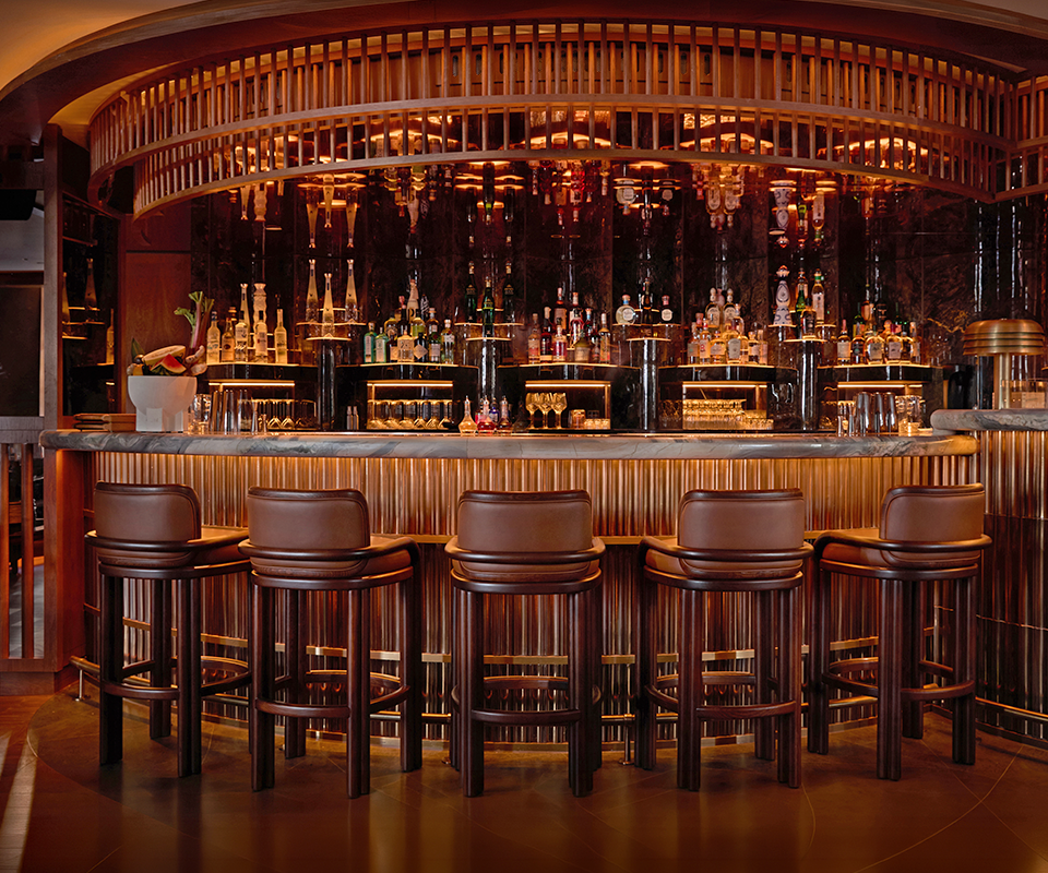 James Bar Chair Row of five James Bar Chairs in smoked walnut wood and tan leather lined along a luxury bar counter in a sophisticated, dimly lit restaurant setting.