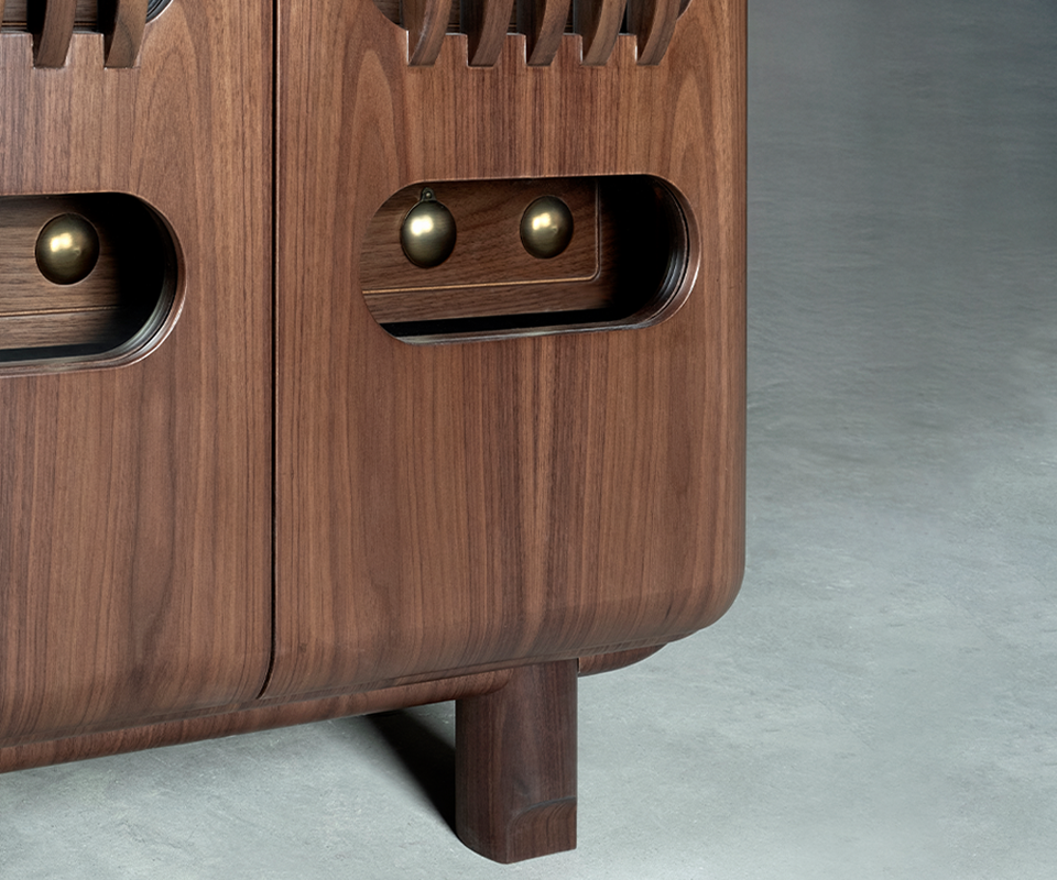A close-up shot of the lower section of the smoked walnut locker. It shows the curved bottom corner, a sturdy square leg, and a recessed handle area on the door featuring spherical details made of aged brushed brass.