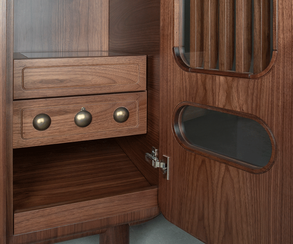 Close-up of the interior lower right corner. It displays a bespoke drawer front in smoked walnut with three spherical aged brushed brass details, positioned next to the open door which contains a glass-protected slatted window.