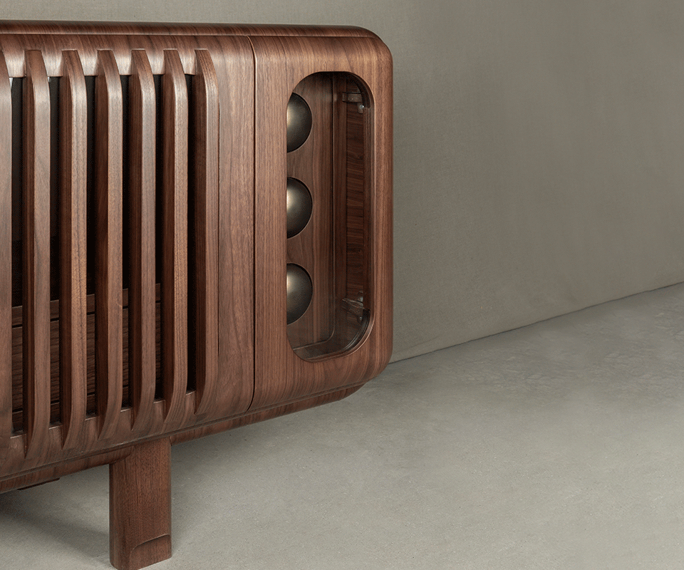 A detailed close-up of the Kennedy Sideboard's corner. The image focuses on the seamless transition between the vertical wooden slats and the glass-fronted end compartment, which houses three metallic brass spheres. The rich wood grain and the rounded silhouette are prominent. The animation showcases the built-in storage.