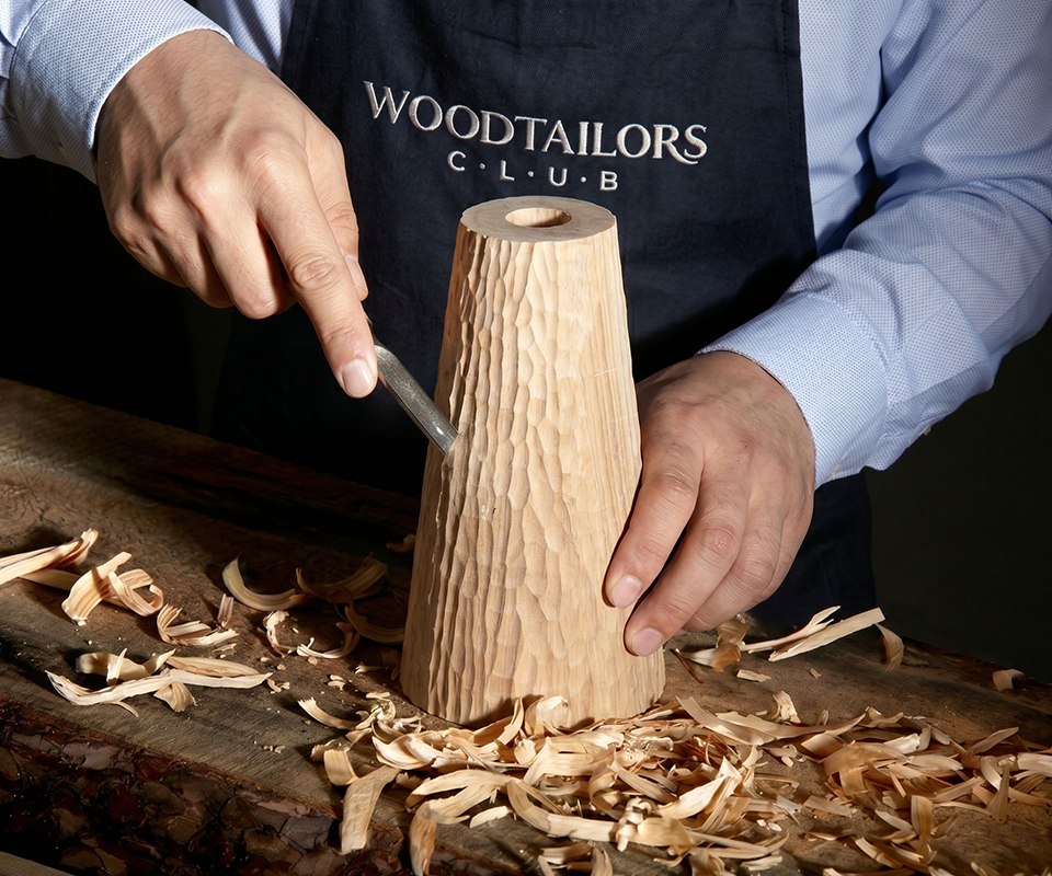 Lloyd I Table Lamp Close-up of a craftsman wearing a Woodtailors Club apron, meticulously hand-carving the textured wooden base of the Lloyd I Table Lamp with a chisel on a workbench surrounded by wood shavings.