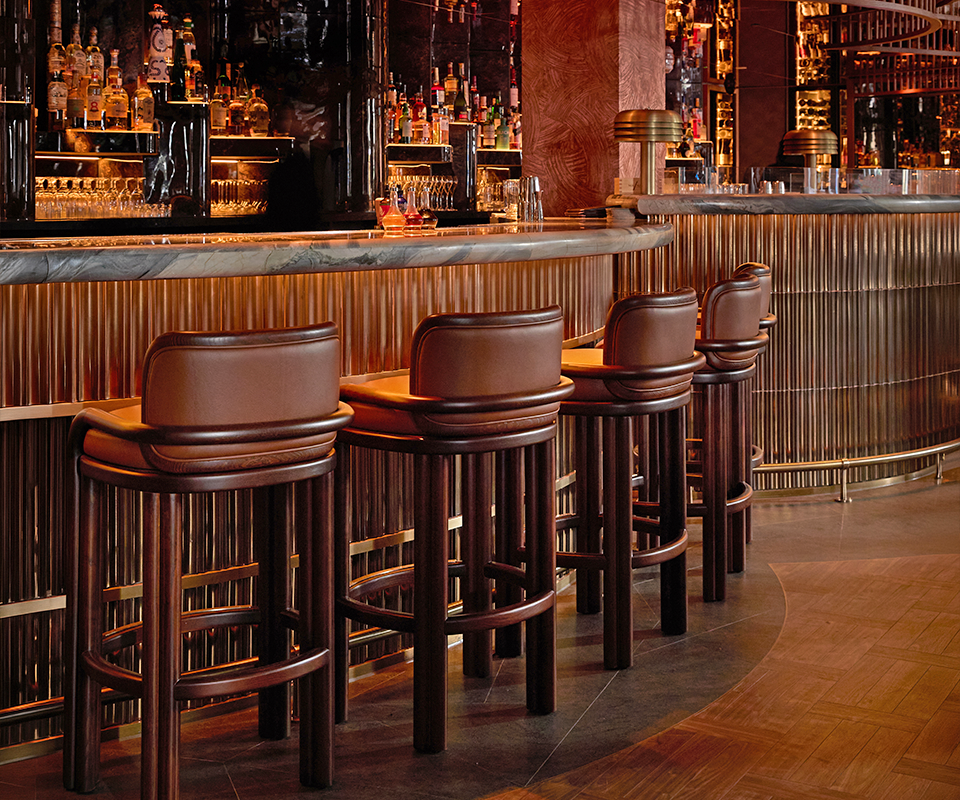 MA/NA Mayfair, London, UK Row of dark wood James Bar Chairs with brown leather upholstery lined up along an elegant, warm-lit marble counter at MA/NA Mayfair, London, UK bar.
