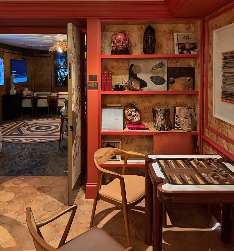 A close-up view of a cozy gaming corner inside The Moore Miami Social Club, showcasing a wooden Holland Backgammon Table set up with checkers and dice. Two modern wooden chairs are placed in front of it. In the background, built-in shelves displaying art books, ceramic masks, and sculptures line a burl wood wall with red trim. An open decorated door reveals another room with a geometric rug and a flat-screen TV in the distance.