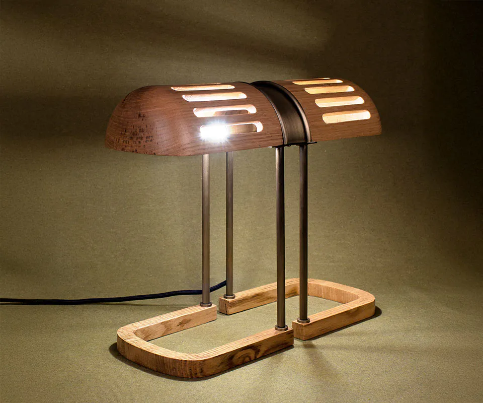 Turing Table Lamp Turing Table Lamp crafted in natural oak wood with aged brass accents, lit with the shade casting beautiful shadows against an olive green background.